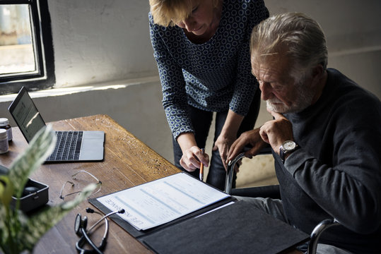 Side View Of Elderly Man Sitting On Wheelchair Looking At Life Insurance Contract Form