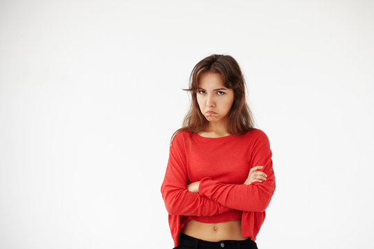 Studio shot of casually dressed young Hispanic female with dark hair pouting, keeping arms folded, looking at camera with unhappy and offended grimace. Negative human expressions and emotions