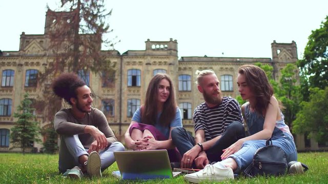 Happy young college fiends sitting on lawn in front of university. College friends preparing for classes together.