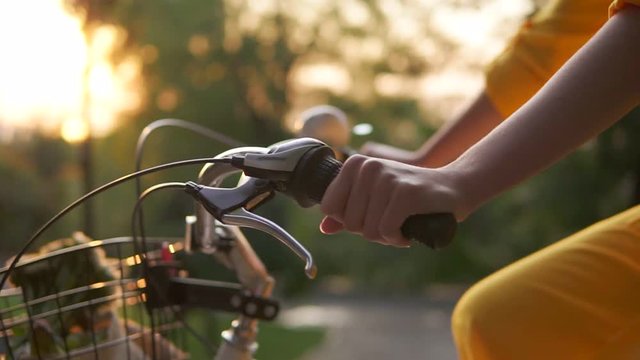 Closeup view of an unrecognizable woman's hands holding a handlebar while riding a city bicycle with a basket and flowers. Lens flare during early moning. Sun is rising. Woman in yellow dress