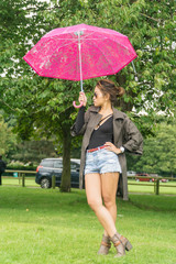 Filipino model under a umbrella walking on a pathway in a park at summertime in the rain, wearing denim shorts a black top with a coat on under a pink umbrella 