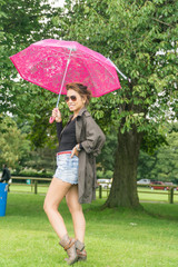 Filipino model under a umbrella walking on a pathway in a park at summertime in the rain, wearing denim shorts a black top with a coat on under a pink umbrella 
