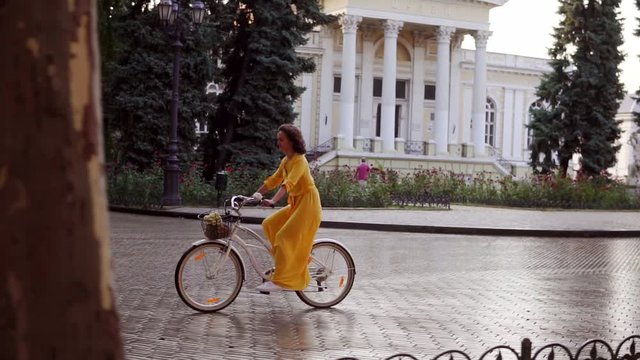 Beautiful woman riding a city bicycle with a basket and flowers in the city center enjoying her time early in the morning. Steadicam shot. Beautiful old city view