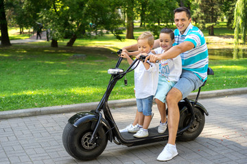 Dad with cute little boys riding electric scooter in summer park outdoors