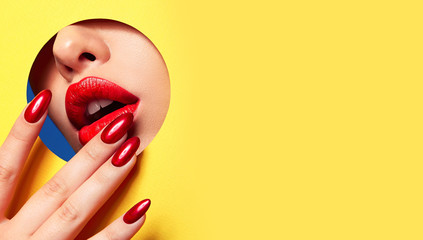 Red lips peep through the hole in the yellow paper. Fingers at the mouth with bright red manicure.