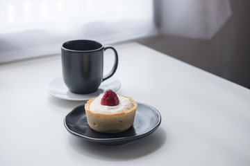 A strawberry cheese tart , a black cup of hot coffee and cactus on white vintage wooden table and curtain background in minimalism cafe
