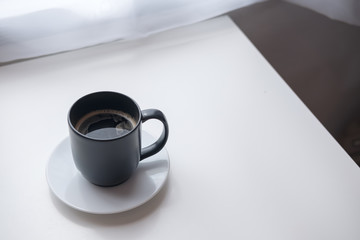Closeup image of a black hot coffee cup with white saucer on vintage wooden table and curtain background in minimalism cafe