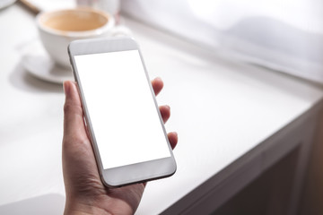Mockup image of a hand holding white mobile phone with blank screen , coffee cup on white vintage wooden table and curtain background in minimalism cafe