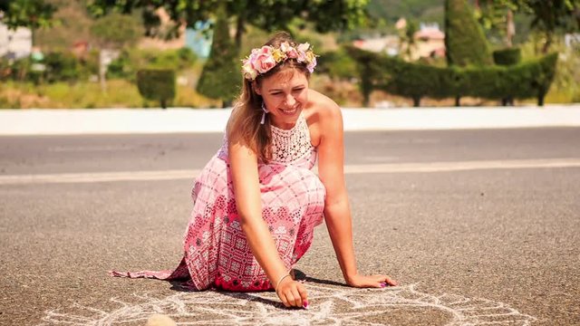Girl in Garland Squats Draws with Chalk on Asphalt Pavement