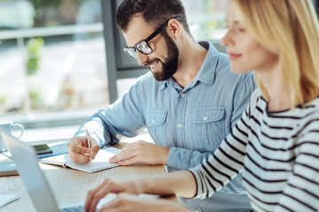 Smiling man writing in notebook while colleague working on laptop