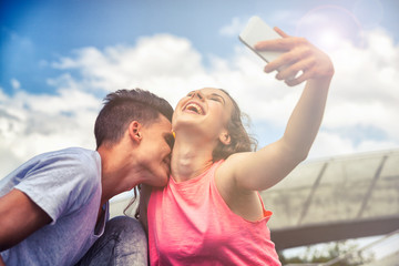 Young couple giving kiss sitting in town outdoor with phone taking selfie