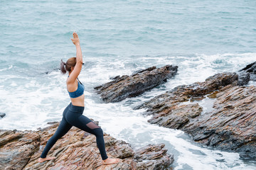 Asian woman standing yoga postures Warrior (virabhadrasana) on the rocks. Sea and sky as background.