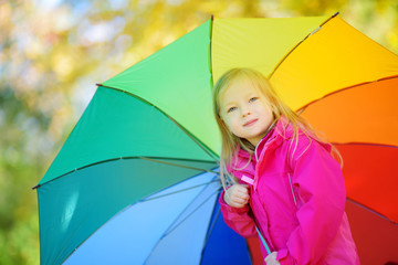 Cute little girl holding rainbow umbrella on beautiful autumn day. Happy child playing in autumn park.