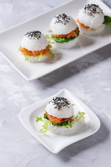 Mini rice sushi burgers with smoked salmon, green salad and sauces, black sesame served on white square plate over gray concrete background. Modern healthy food