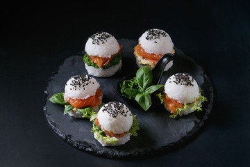 Mini rice sushi burgers with smoked salmon, green salad and sauces, black sesame served on slate stone board over black background. Modern healthy food