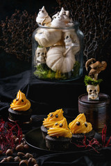 Halloween decorated sweet dessert table black cupcake with orange cream, white meringue ghosts with chocolate eyes on plate, decor skulls and pumpkin over black tablecloth.
