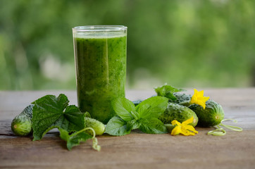  glass with green smoothie on a wooden table and  cucumbers with flowers and mint leaves