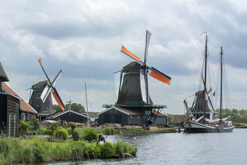 Three dutch mills with orange wings near the canal with one beautiful boat under the cloudy sky in Zaanse Schans (Netherlands)