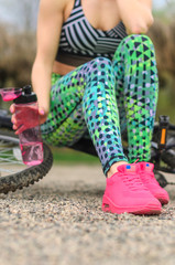 Obraz premium brunette girl in sportswear, riding a Bicycle in the morning in the Park. Resting and drinking water .Closeup of the foot