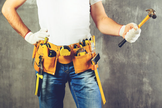 unidentified builder standing in white gloves with a tool belt with construction tools and holding a hammer against grey background, toned image. DIY tools and manual work concept.