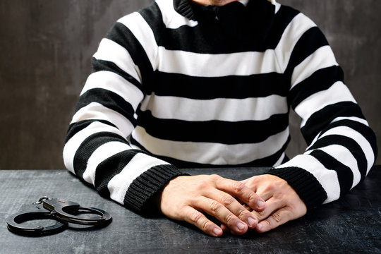 unidentified prisoner in prison stripped uniform sitting on the chair in the dark interrogation room, next to him handcuffs laying on the table. The interrogation of the arrested