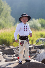 Smiling child boy in traditinal Polish mountain outfit and black hat stands on mountain rocks - obrazy, fototapety, plakaty