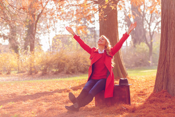 Beautiful young woman siting on vintage suitcase in a autumn park with her arms outstretched.