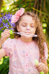 Happy little curly girl playing with soap bubbles on a summer nature, wearing a pink dress and flower in her head, in a blurred nature background