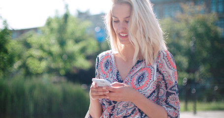 Young blonde girl using phone, outdoors. Beautiful young woman typing on phone in a city during sunny day.