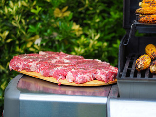 Raw beef steaks on cutting board ready for barbecue and roasted corn on grill.