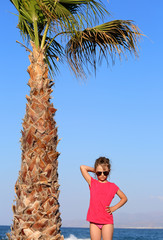 Beautiful cute kid gril in fashion trendy sunglasses posing near the palm on blue sky and sea background