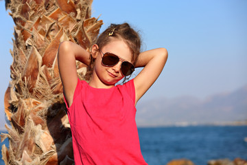 Cute beautiful kid girl in fashion sunglasses standing and relaxing near the palm on blue sea and sky background