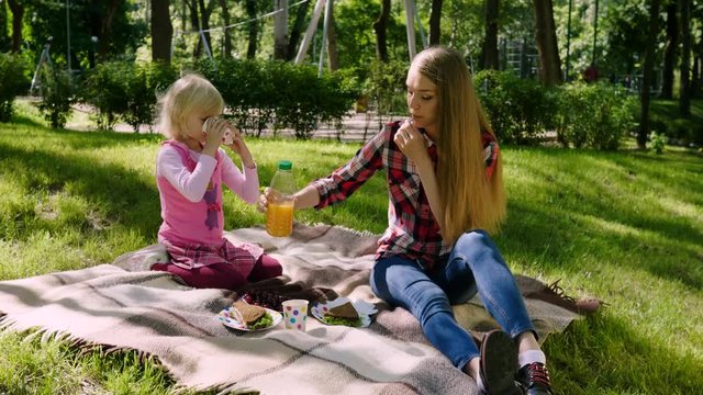 Picnic outdoors. The young blond woman pouring juice in a paper cup to a little cheerful girl. 4K