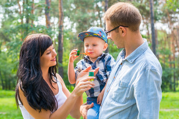 A little boy in the hands of the dad and blow soap bubbles. Mom stands by and smiles.