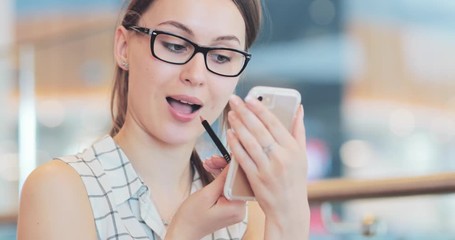 Young Woman Paint Her Lips, using Smartphone as Mirror. 4K DCi SLOW MOTION, CLOSE UP. Stylish Girl using lipstick in the shopping mall. Cinematic Shot