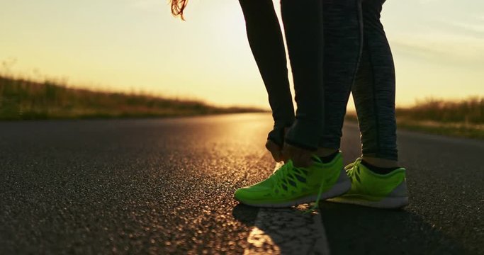 Female Athlete Tying Laces, Getting Ready for Training. SLOW MOTION 4K DCi. Runner preparing for jogging on an open countryside road. Morning practice at sunrise. Cinematic sports shot. 