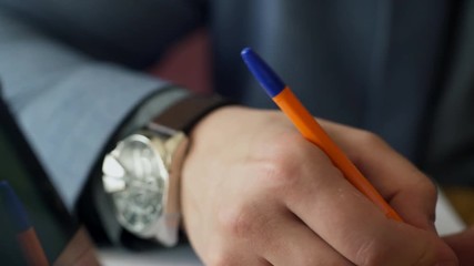 Man's hand with expensive watches on his wrist writes pen on paper. Business man quickly takes notes in his notebook during the meeting. A businessman in a gray suit outlines information in orange - Powered by Adobe