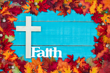 Religious cross and the word Faith hanging on wooden teal blue background with colorful autumn leaves border