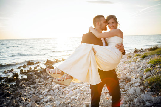 Groom carrying bride on pebble beach at sunset