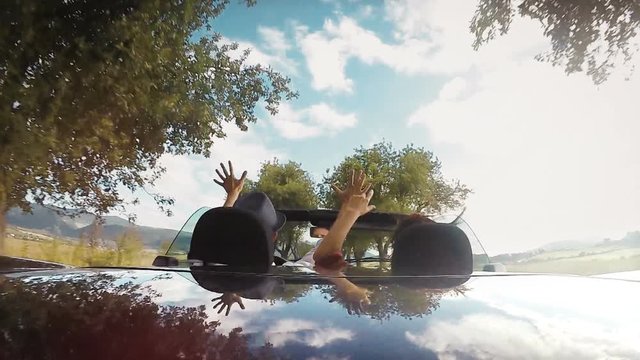 Slow-motion footage of two people driving in the convertible car and raising their hands