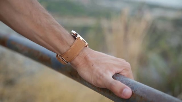 Mano di un uomo che si appoggia a una ringhiera con un orologio marrone al polso e uno sfondo di campagna sfuocato. 