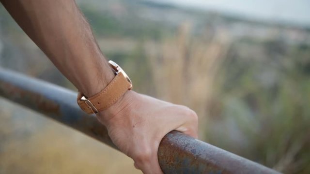 Mano di un uomo che si appoggia a una ringhiera con un orologio marrone al polso e uno sfondo di campagna sfuocato. 
