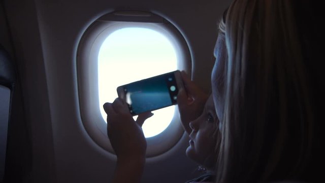 Mother and daughter taking shots with smartphone through illuminator in airplane.