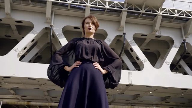 Steadicam shot. the girl middle-aged with contrasting makeup in dark loose clothing on the background of bridge rail in a modern city