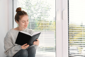 Beautiful young girl sitting on window sill with book at home