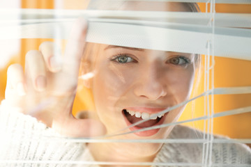 Beautiful young girl separating slats of blinds and looking through window
