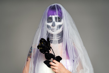 Portrait of woman with terrifying halloween skeleton makeup and purple wig bridal veil, wedding dress holds a black rose in his hands over gray background. Black wedding
