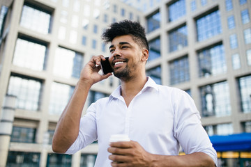 Young handsome Afro American man standing in front of huge modern business building smiling and talking on cell phone