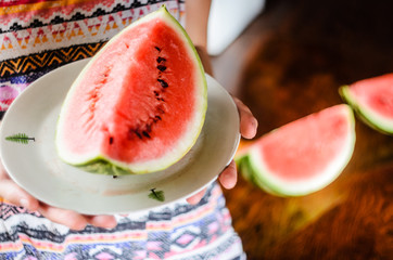 Watermelon background. Girl in a colorful summer shirt holding a plate with a large slice of watermelon against a background of a blurry table with slices of watermelon