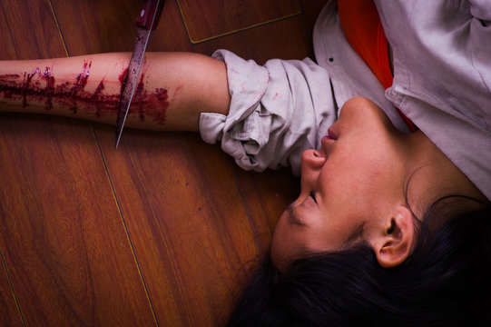Close up of a young depressive woman holding a knife, with her arm bleeding, laying on the ground, in a wooden background